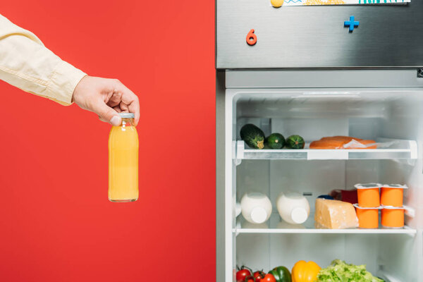 cropped view of man holding bottle of juice near open fridge with fresh food on shelves isolated on red