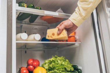 Buzdolabından peyniri alıp raflarda taze yiyecek taşıyan bir adamın görüntüsü.