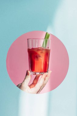 cropped view of woman holding bloody Mary with celery in pink circle on blue background in sunlight clipart