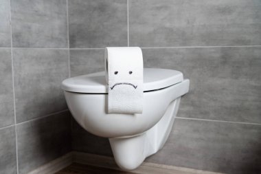 Toilet paper with sad emoticon on toilet bowl in modern restroom clipart