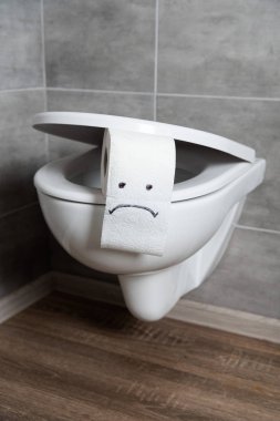 Sad emoticon on white toilet paper on toilet bowl in modern restroom clipart