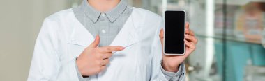Cropped view of pharmacist pointing with finger at smartphone with blank screen, panoramic shot clipart