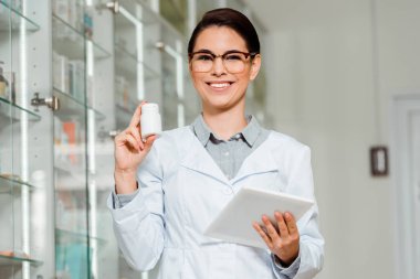 Güzel eczacı kameraya gülümserken elinde bir kavanoz hap ve dijital tablet tutuyor. 