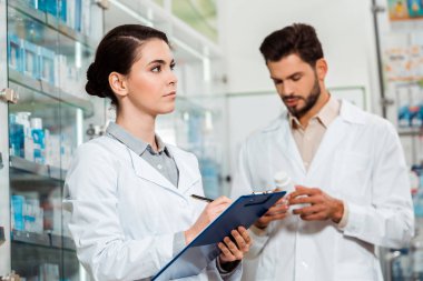 Pensive pharmacist writing on clipboard and colleague with pills on background clipart