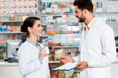 Eczacıların eczanede birbirlerine gülümseyen ilaç ve not panosuyla yan bakışları