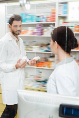 Selective focus of pharmacist with pills looking at colleague at drugstore counter clipart