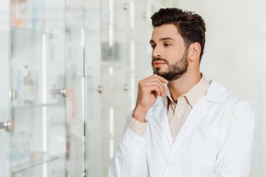 Pensive druggist looking at showcase shelves in pharmacy clipart