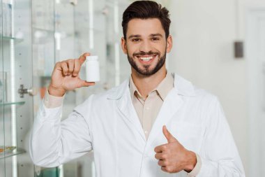Pharmacist with jar of pills smiling at camera and showing thumb up  clipart