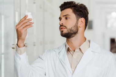Eczanedeki beyaz önlüklü eczacı kavanoza haplarla bakıyor.