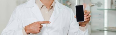 Cropped view of pharmacist in white coat pointing at smartphone with blank screen, panoramic shot clipart