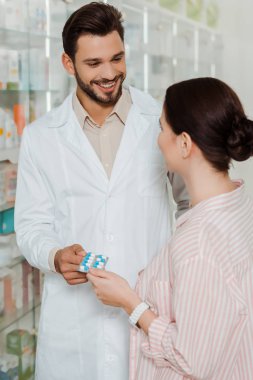 Smiling druggist giving blister with pills to customer by pharmacy showcase clipart