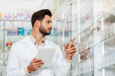 Handsome pharmacist with digital tablet pointing at showcase in pharmacy  clipart