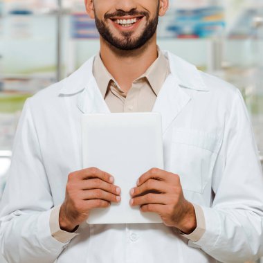 Beyaz önlüklü, elinde dijital tablet olan gülümseyen eczacı görüntüsü.