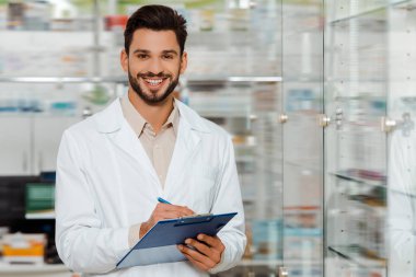 Handsome pharmacist with clipboard smiling at camera by drugstore showcase clipart