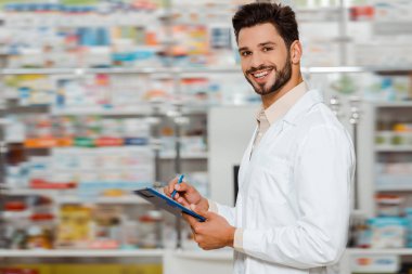 Selective focus of smiling pharmacist with clipboard looking at camera in apothecary clipart