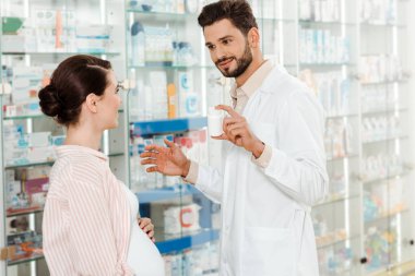 Smiling pharmacist showing to pregnant woman jar with pills by drugstore showcase clipart