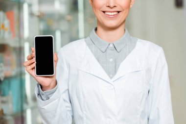 Gülümseyen eczacının elinde boş ekran ile akıllı telefon tutmasının kırpılmış görüntüsü 