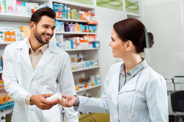 Smiling pharmacist giving to colleague jar with pills in pharmacy