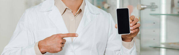 Обрезанный вид аптекаря в белом халате, указывающий на смартфон с пустым экраном, панорамный снимок
