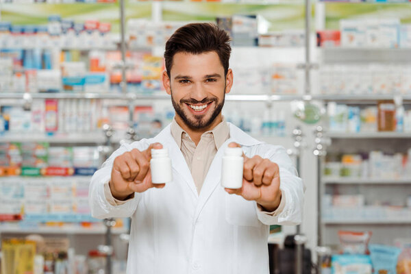 Улыбающийся аптекарь смотрит в камеру и держит банки с таблетками
