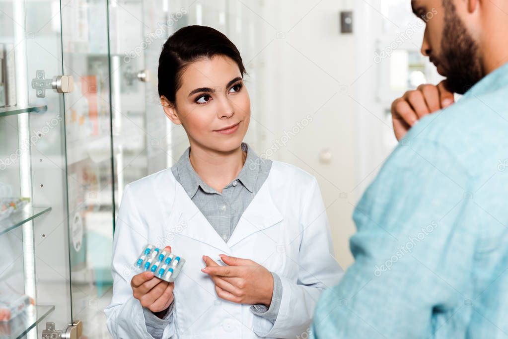 Selective focus of pharmacist holding blister with pills and looking at customer 