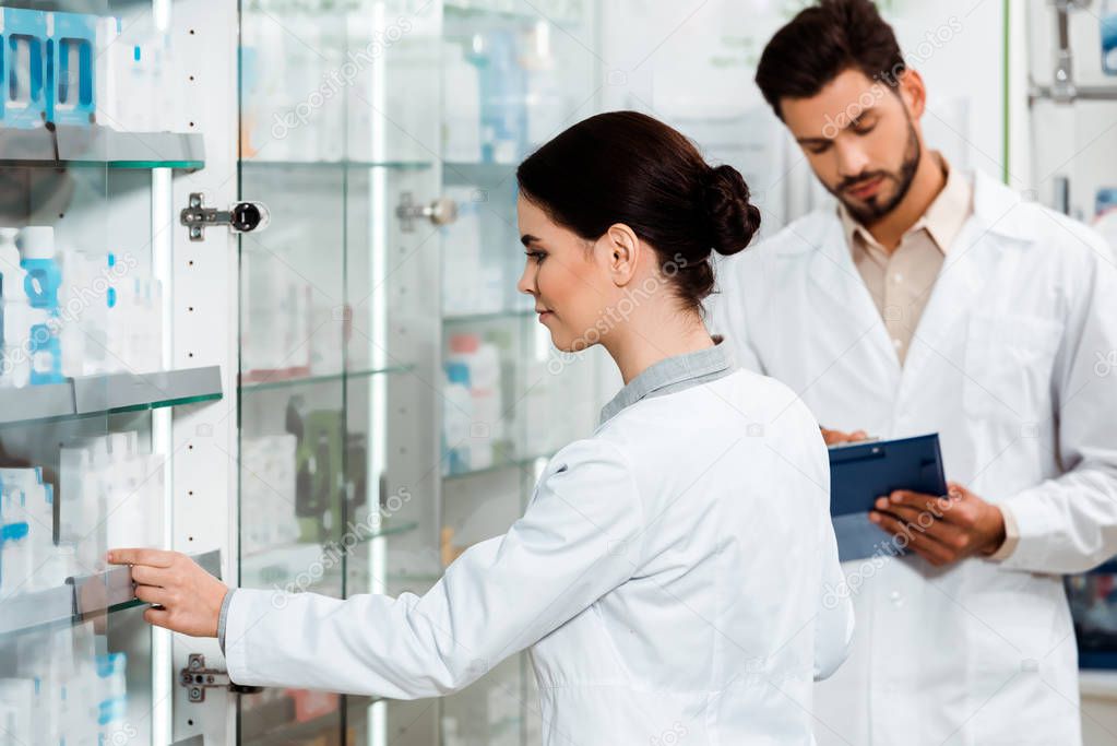Pharmacist taking medicament from showcase beside colleague with clipboard in pharmacy