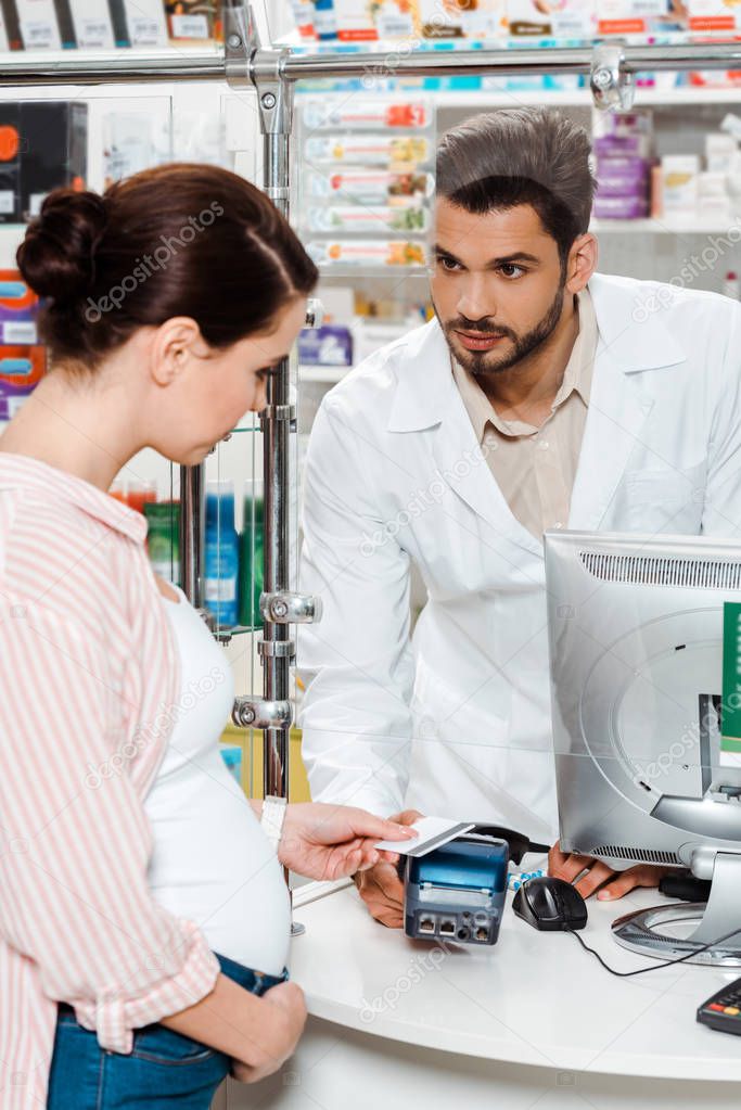 Pharmacist looking at pregnant woman paying with paypass in drugstore