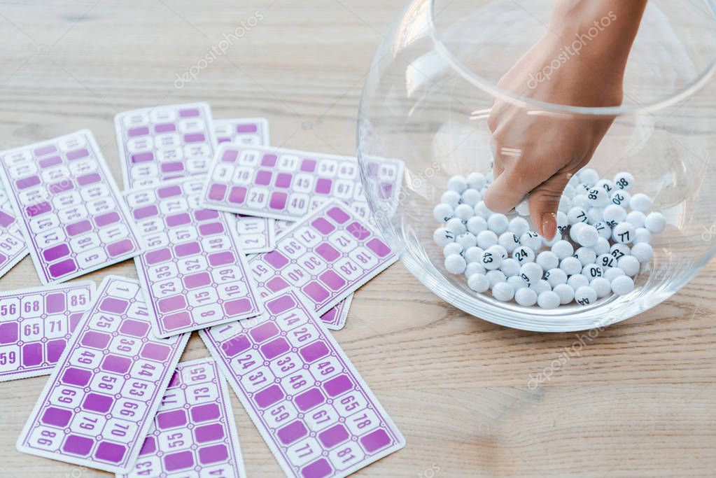 cropped view of woman taking ball from glass bowl near lottery tickets 