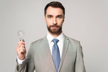 confident businessman holding light bulb and looking at camera isolated on grey clipart