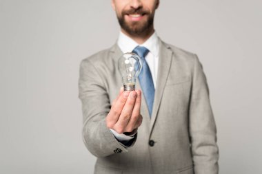 partial view of smiling businessman holding light bulb isolated on grey clipart