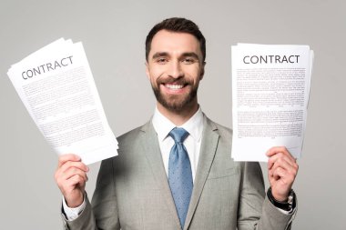 Sözleşmeleri tutan mutlu iş adamı gri kameraya bakıyor.