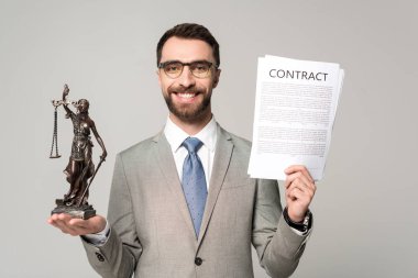 handsome lawyer holding contract and themis statue while smiling at camera isolated on grey clipart