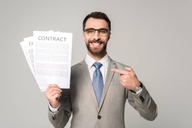 smiling businessman in glasses pointing with finger at contracts isolated on grey clipart
