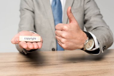 cropped view of businessman holding wooden block with word copyright and showing thumb up isolated on grey clipart