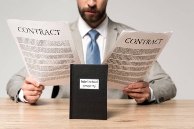 cropped view of businessman reading contracts near book with intellectual property title isolated on grey clipart