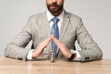 partial view of businessman showing protection gesture above light bulb isolated on grey clipart