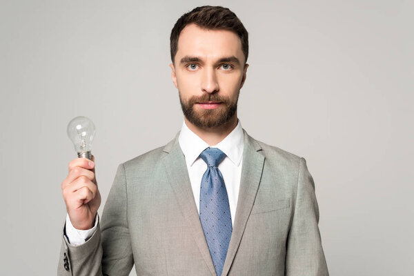 confident businessman holding light bulb and looking at camera isolated on grey