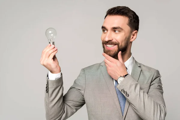 Lächelnder Geschäftsmann Berührt Gesicht Während Glühbirne Isoliert Auf Grau Hält — Stockfoto