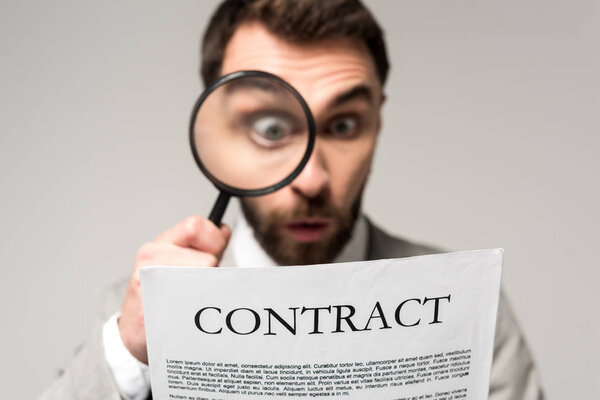 selective focus of shocked businessman looking at contract through magnifier isolated on grey