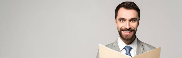 панорамный снимок позитивного бизнесмена, улыбающегося в камеру, держащего в руках бумажную папку, изолированную на сером
