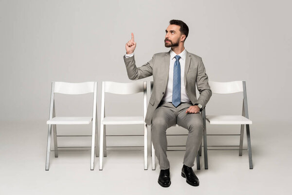 confident businessman sitting on chair and showing idea gesture on grey background