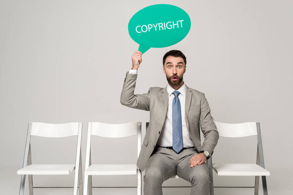 surprised businessman holding thought bubble with word copyright while sitting on chair on grey background