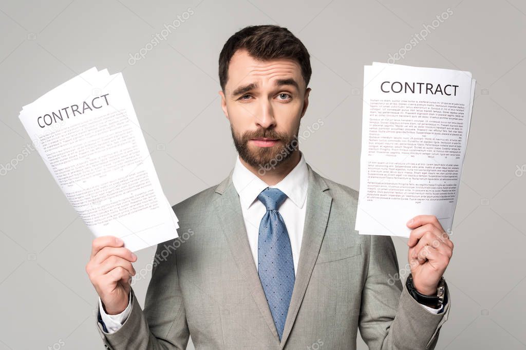 skeptical businessman holding contracts and looking at camera isolated on grey