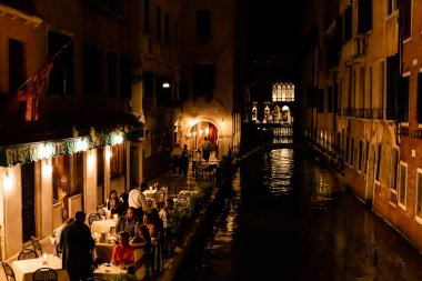 Venedik, İtalya - 24 Eylül 2019: Venedik, İtalya 'da geceleri kanal manzaralı açık kafe yakınında oturan turistler 