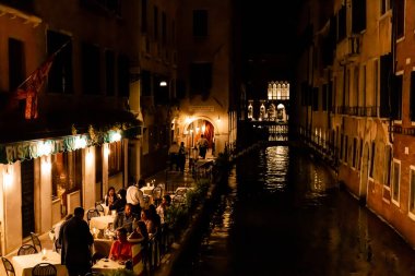 VENICE, ITALY - SEPTEMBER 24, 2019: tourists sitting near outdoor cafe with view at canal at night in Venice, Italy  clipart