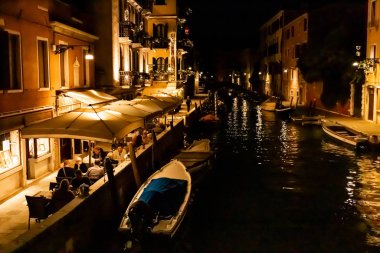 Venedik, İtalya - 24 Eylül 2019: Venedik, İtalya 'da geceleri kanal manzaralı açık kafe yakınında oturan turistler 
