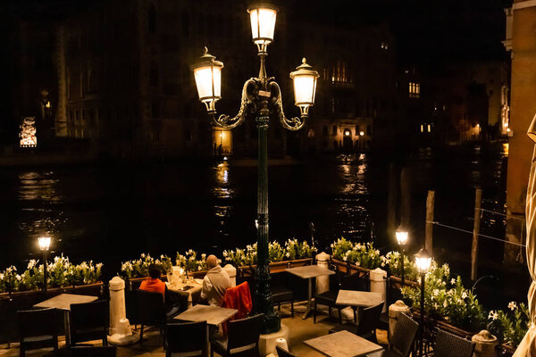 VENICE, ITALY - SEPTEMBER 24, 2019: tourists sitting at table and looking at canal at night in Venice, Italy 