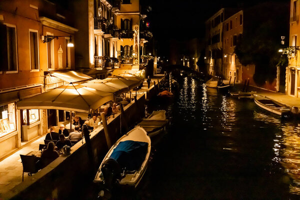 VENICE, ITALY - SEPTEMBER 24, 2019: tourists sitting near outdoor cafe with view at canal at night in Venice, Italy 