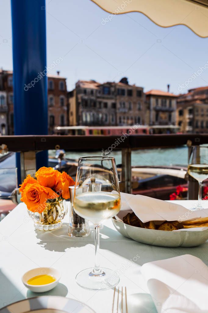 selective focus of wine glass, bread and flowers on table and ancient buildings on background 