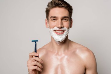 smiling sexy man with muscular torso and shaving foam on face holding shaver isolated on grey clipart
